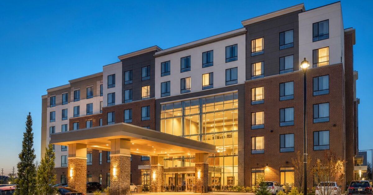 Exterior of a modern upper-midscale all-suite hotel at dusk with illuminated lobby entrance and surface parking